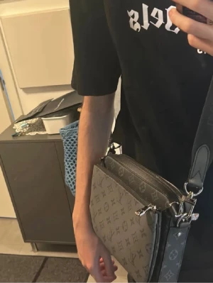 Louis Vuitton axelväska monogram grå - Snygg axelväska från Louis Vuitton i grått och svart med klassiskt monogrammönster. Väskan har justerbar axelrem och detaljer i svart läder. Perfekt för dig som vill ha en stilren och lyxig accessoar till din outfit.Perfekt för dig som vill resa och inte ha allt i fickorna 