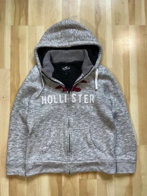 Grå zip hoodie från Hollister - Snygg gråmelerad hoodie från Hollister med dragkedja, huva och vita dragsnören. Stor logga i vitt och rött broderi på bröstet. Mjuk insida och två fickor framtill. Perfekt för en avslappnad och cool stil.