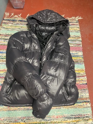 Svart pufferjacka från Brave Soul - Säljer en svart pufferjacka från Brave Soul med glansig finish och huva. Jackan har dragkedja framtill, två sidofickor med dragkedja och quiltad design. Perfekt för kalla dagar och riktigt skön att bära.