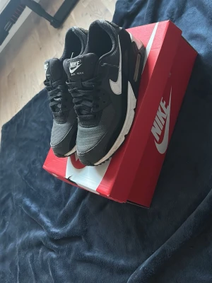 Nike Air Max 90 svarta sneakers - Nike Air Max 90 sneakers i svart med vita detaljer och klassisk synlig Air-sula. Ovandelen är i mesh och läder med vita swoosh-loggor på sidorna och AIR-tryck bak. Snygg retrostil som passar till streetwear och casual outfits.