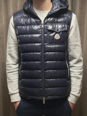 Mörkblå dunväst från Moncler - Snygg mörkblå dunväst från Moncler med glansig finish och horisontella sömmar. Västen har huva, bröstficka med lock och Moncler-logga samt dragkedja framtill. Perfekt för lager på lager och streetwear-vibe.