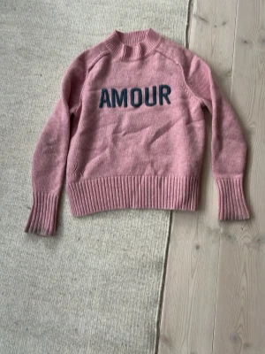 Rosa stickad tröja med text från Zadig Voltaire  - Säljer en rosa stickad tröja med mörkblå texten 'AMOUR' på bröstet. Tröjan har ribbade muddar vid ärmslut och nederkant samt hög hals. Perfekt för dig som gillar en trendig och mysig look. Tröjan är från Zadig Voltaire i storlek 12 år/xs