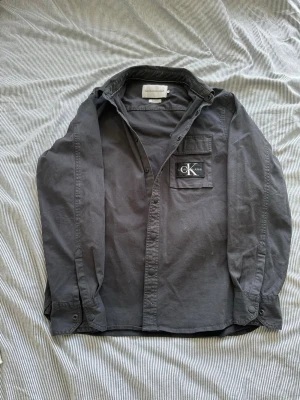 Mörkgrå skjorta från Calvin Klein - Snygg mörkgrå skjorta från Calvin Klein Jeans med klassisk krage och knappstängning. Bröstficka med stor CK-logga som detalj. Perfekt för en clean och stilren look.