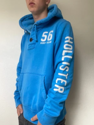 Blå hoodie från Hollister - Fett hoodie från hollister. Modellen är 178cm och väger cirka 70kg. Hoodien har ett tryck vid armen där det står ”Hollister”. Hör gärna av er vid intresse eller frågor!