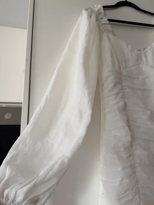 Vit kort klänning med puffärm - Säljer en vit kort klänning med långa puffärmar och v-ringad hals. Klänningen har ett lätt rynkat tyg och volangkant nedtill. Perfekt för dig som vill ha en romantisk och trendig look. Materialet känns som tunn polyester eller liknande. Oanvänd!