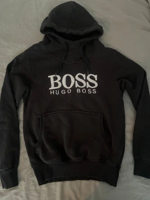 Svart hoodie från Hugo Boss - Snygg svart hoodie från Hugo Boss med stor vit logga på bröstet. Hoodien har huva med dragsko, magficka och ribbade muddar vid ärmar och nederkant. Tillverkad i mjukt material som känns skönt mot huden. Perfekt för en avslappnad och trendig look.