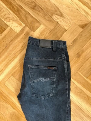 Mörkblå jeans från Nudie Jeans - Säljer ett par mörkblå jeans från Nudie Jeans med klassisk femficksmodell och raka ben. Jeansen har snygga slitningar framtill och kontrastsömmar på bakfickan. Tillverkade i denim med en skön passform. Perfekta för en avslappnad stil. Total längd 91cm
