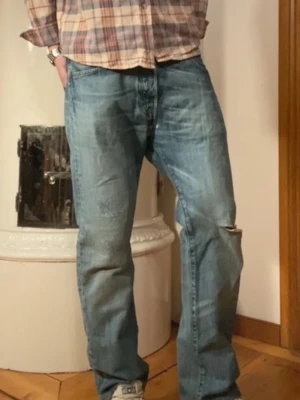 Blå raka jeans från Levi's - Säljer ett par klassiska blå jeans från Levi's med rak passform och slitningar på vänster knä. Jeansen har fem fickor, gylf med dragkedja och knapp samt typisk Levi's-design. Perfekta för en avslappnad och trendig stil.