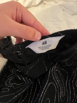 Svart topp med silvermönster från H&M - Snygg svart topp från H&M med glittrigt silvermönster och knyt framtill. Toppen har ett unikt abstrakt mönster och är tillverkad i ett mjukt syntetmaterial. Perfekt för dig som vill sticka ut lite extra.