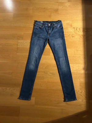 Jack and Jones jeans - Säljer ett par blå skinny jeans med lätt slitna detaljer och klassisk femficksdesign. Jeansen har normalhög midja och smala ben. Materialet är stretchigt denim som sitter bekvämt. Perfekta till sneakers och sommaren
