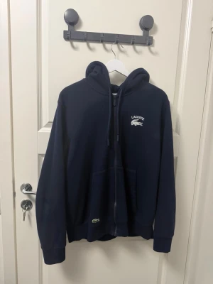 Mörkblå hoodie från Lacoste - Snygg mörkblå hoodie från Lacoste med dragkedja framtill och klassisk krokodil-logga broderad på bröstet. Hoodien har huva med dragsko och två stora fickor framtill. Tillverkad i mjukt bomullsmaterial som känns skönt mot huden. Perfekt för en chill och sportig look.