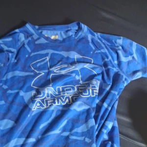 Blå camo t-shirt från Under Armour - Snygg blå t-shirt från Under Armour med camouflagemönster och stor logga på bröstet. Tillverkad i ett lätt och snabbtorkande material som passar perfekt till träning eller sportaktiviteter. Rund halsringning och korta ärmar.