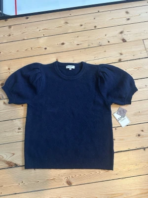 Mörkblå puffärmströja Samsa Young - Mörkblå stickad tröja från Samsa Young med korta puffärmar och rund halsringning. Tröjan har ribbade muddar vid ärmslut och nederkant, vilket ger en snygg och modern look. Perfekt för dig som gillar trendiga detaljer och enkel stil.