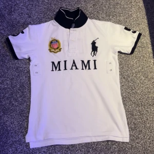 Vit pikétröja från Ralph Lauren - Säljer en snygg vit pikétröja från Ralph Lauren med svart krage och broderad logga. Tröjan har texten 'MIAMI' på framsidan och siffran '5' på baksidan. Perfekt för en sportig look!