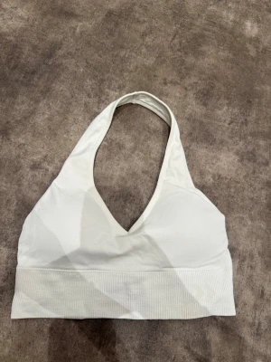 Vit halterneck sport-bh - Säljer en vit halterneck sport-bh med bred ribbad kant nedtill och diskret logga framtill. Bh:n har en djup v-ringning och är tillverkad i ett stretchigt material. Öppen i ryggen. Skulle säga att det är en medium även om det står smal. 