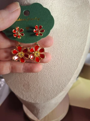 Röda blommiga örhängen i metall - Snygga örhängen i form av röda blommor med gulddetaljer och glansig finish. Blommorna har flera kronblad och en gul mitt, vilket ger ett färgstarkt och lekfullt intryck. Perfekta för att lyfta vilken outfit som helst.
