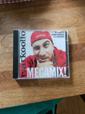 Markoolio Megamix CD - CD-skiva med Markoolio Megamix! Omslaget är rött och vitt med artistens namn och låttitlar som 'Gör det igen', 'Vi drar till fjällen', 'Sola & bada' och fler. Plastfodral med tryckt framsida och baksida.