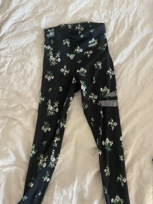 Svarta träningstights med blommor Stronger - Snygga svarta träningstights från Stronger med blommigt mönster i grönt, vitt och lila. Hög midja och stretchigt material som sitter skönt vid träning. Perfekta för gymmet eller löprundan.