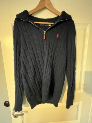 Mörkblå kabelstickad tröja Polo Ralph Lauren - Mörkblå kabelstickad tröja från Polo Ralph Lauren med halv dragkedja och hög krage. Tröjan har ett rött broderat logomärke på bröstet och ribbade muddar vid ärmslut och nederkant. Perfekt för lager på lager och klassisk stil.