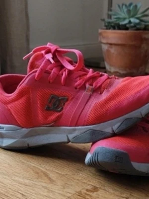 Rosa DC sneakers med meshdetaljer - Snygga sneakers från DC i stark rosa färg med meshmaterial för bra ventilation. Skorna har låga skaft, vita och grå detaljer på sulan samt klassisk DC-logga på sidan. Perfekta för dig som vill sticka ut med färg och sportig stil.
