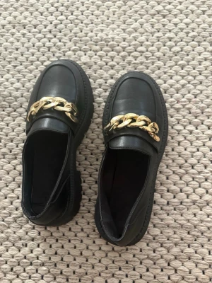 Svarta chunky loafers med guldkedja - Säljer ett par svarta loafers med chunky sula och en stor guldfärgad kedjedetalj över foten. Skorna har en stilren design och är tillverkade i ett slätt material som liknar läder. Perfekta för dig som vill ha en trendig och edgy look.