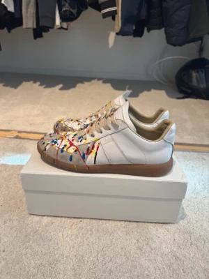 Maison Margiela sneakers med färgstänk - Unika vita sneakers från Maison Margiela med färgstänk i blått, rött, gult och svart. Skorna har klassisk låg siluett, vita snören och beige sula. Tillverkade i läder och levereras i originalkartong med dustbags. Perfekt för dig som vill sticka ut med din stil.