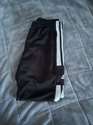 Svarta Adidas träningsbyxor - Säljer ett par svarta träningsbyxor från Adidas med klassiska vita ränder längs sidorna. Byxorna har resår i midjan och är tillverkade i ett glansigt, lätt material som passar perfekt för sport och träning.