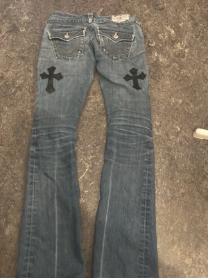Blå bootcut jeans med svarta kors - Unika blå bootcut jeans med stora svarta korsapplikationer på både fram- och baksida. Jeansen har klassiska fem fickor, kontrastsömmar och lätt slitna detaljer. Perfekt för dig som vill sticka ut med en edgy och personlig stil men man behöver bara fixa till gylfen lite.