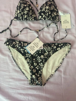 Svart blommig bikini från H&M - Snygg svart bikini från H&M med vita små blommor och tunna knytband. Både överdel och underdel har ett blommigt mönster och bikinin är tillverkad av återvunnen polyamid och polyester. Perfekt för strandhäng eller poolparty!