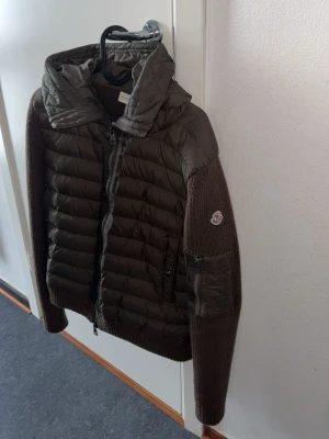 Mörkbrun pufferjacka från Moncler - Snygg mörkgrön pufferjacka från Moncler med quiltad design och ribbstickade ärmar. Jackan har dragkedja framtill, fickor med knapp och en extra ficka med dragkedja på ärmen. Perfekt för kalla dagar och har Moncler-logga på ärmen. Finns att hämta i ÖREBRO kan också fraktas.