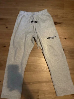 Grå Essentials mjukisbyxor - Snygga ljusgrå helt nya mjukisbyxor från Essentials med tryck på vänster lår. Byxorna har resår i midjan med dragsko och raka ben. Perfekta för chill dagar och streetwear-stil. Mjukt material som känns skönt mot huden.
