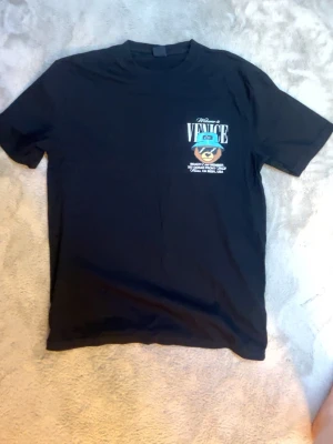 Svart t-shirt med björntryck Venice - Cool svart t-shirt med stort tryck på ryggen av en björn med blå hatt och texten 'Welcome to Venice'. Mindre tryck på bröstet. Perfekt för dig som gillar streetwear och grafiska motiv.