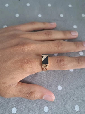 Guldfärgad ring med svart sten - Snygg guldfärgad ring med en rektangulär svart sten och geometriska detaljer på sidorna. Ringen har en modern design med öppna partier och små glittrande stenar i ett hörn. Perfekt för dig som gillar statement-smycken.