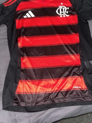 Randig fotbollströja från Adidas Flamengo  - Snygg fotbollströja från Adidas i slim fit-modell. Tröjan har breda röda och svarta horisontella ränder, V-ringad krage och är tillverkad i ett lätt och ventilerande funktionsmaterial. Perfekt för träning eller match.