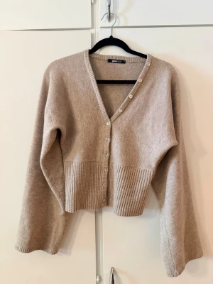 Beige kofta med v-ringning från ginatricot - Beige kofta från ginatricot med v-ringning och pärlemorknappar framtill. Modellen är croppad med bred ribbstickad kant nedtill och har långa, lite vidare ärmar. Mjuk och skön stickning som passar perfekt till jeans eller kjol.