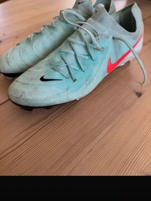 Nike phantom gx 2 - Säljer ett par Nike fotbollsskor i ljusblå färg med klassisk snörning och röda detaljer på sidan. Skorna har en låg profil och dobbar för bra grepp på planen. Perfekta för dig som vill ha stilrena och funktionella skor till fotbollsträningen.