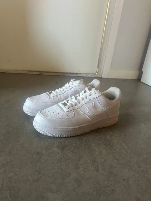 Nike Air Force 1 vita sneakers - Klassiska helvita Nike Air Force 1 sneakers i lågt utförande. Skorna har en stilren design med perforerad tå, kraftig sula och ikoniska swoosh-loggan på sidan. Tillverkade i läder med vita skosnören och diskret branding på hälen och sulan.