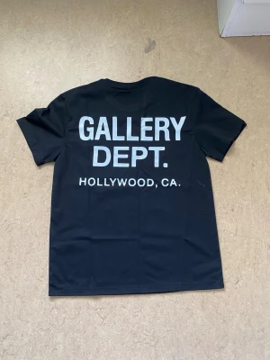 Svart t-shirt från Gallery Dept. - Svart t-shirt från Gallery Dept. med vit text på både bröstet och ryggen. Klassisk rund halsringning och korta ärmar. Stort tryck på ryggen med 'GALLERY DEPT. HOLLYWOOD, CA.' och mindre logga fram. Perfekt för en streetwear-look.