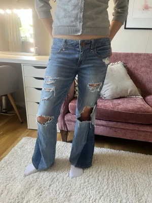 Blå slitna bootcut jeans - Säljer ett par blå bootcut jeans med stora slitningar och hål på knäna och låren. Jeansen har klassisk femficksmodell, låg midja och råa kanter nertill. Perfekta för en avslappnad och trendig look.