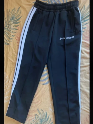 Palm Angels svarta byxor  - Säljer ett par svarta trackpants från Palm Angels med vita sidoränder och logga på vänster ben. Byxorna har resår i midjan och raka ben. Perfekta för en sportig och avslappnad stil. Materialet är troligtvis polyester för en mjuk och bekväm känsla.