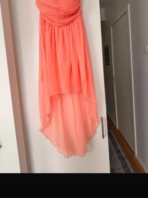 Korallfärgad axelbandslös klänning - Säljer en snygg korallfärgad klänning med omlott över bysten och asymmetrisk nederkant. Klänningen är axelbandslös och har dragkedja i ryggen. Perfekt för sommarens alla tillfällen!