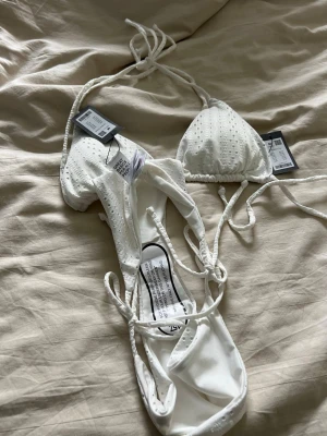Vit bikini med knyt från Lager 157  - Snygg vit bikini från lager 157  med triangeltopp och knyt i sidorna samt nacken. Bikinin har diskreta små hål som ger en broderad känsla och är tillverkad i mjuk syntet. Perfekt för lata dagar på stranden eller poolen.