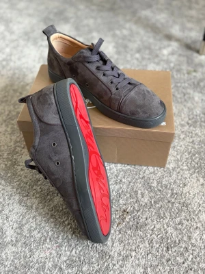 Gråa Louboutin sneakers i mocka - Snygga gråa sneakers från Christian Louboutin i mjuk mocka med klassisk röd sula och svart yttersula. Lågt skaft och stilren design med matchande grå skosnören. Perfekta för dig som vill ha något exklusivt och trendigt i garderoben.