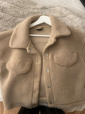 Beige teddyjacka med fickor - Mysig beige teddyjacka med krage och två bröstfickor. Jackan har knappstängning framtill och är gjord i ett fluffigt, mjukt material som ger en avslappnad och trendig look. Perfekt för kyliga dagar när du vill vara både varm och snygg.
