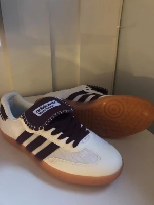 Adidas Wales Bonner sneakers vit/lila - Säljer ett par Adidas Wales Bonner sneakers med vit ovandel, lila detaljer och klassiska tre ränder. Skorna har grov sula i gummi och kontrastsömmar runt plösen. Materialet är en mix av mocka och textil. Perfekta för dig som gillar unika samarbeten och retrostil.