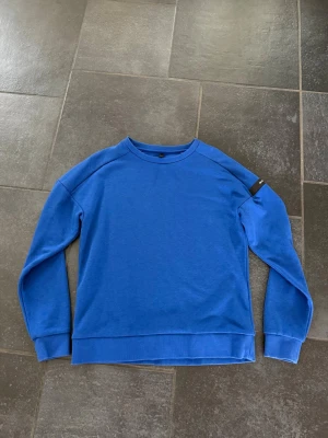 Sail Racing  - Blå  tjocktröja från Sail Racing med rund halsringning och ribbade muddar. Tröjan har en diskret logga på ärmen och en liten logga i nacken. Perfekt för en avslappnad och stilren look.