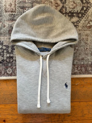 Ljusgrå Polo Ralph Lauren Hoodie - Storlek M - Hej! 👋 Säljer en klassisk ljusgrå Polo Ralph Lauren hoodie i storlek M med marinblå pony-logga. 🐎✨ Tröjan är i riktigt fint skick, välvårdad och helt utan hål eller fläckar. Superfräsch! 100% äkta med alla originaltvättlappar kvar (ingen QR-kod på denna årsmodell). Det mest mångsidiga premium-basplagget man kan ha i garderoben – passar till allt! Fraktar snabbt! 🚛📦💬 