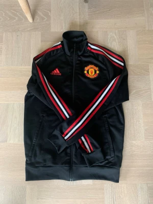 Manchester United träningsjacka Adidas - Snygg svart träningsjacka från Adidas med Manchester Uniteds klubbmärke på bröstet. Jackan har röda och vita ränder längs ärmarna, hög krage och dragkedja framtill. Perfekt för fotbollsträning eller som sportig streetwear. Fint skick använd fåtal gånger, storlek S.