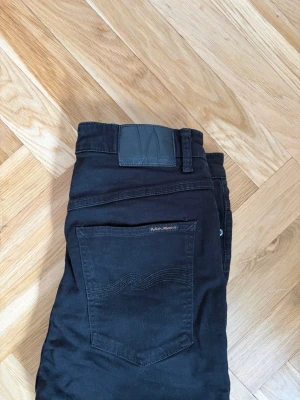 Svarta skinny jeans från Nudie Jeans - Säljer ett par svarta skinny jeans från Nudie Jeans med klassisk femficksdesign och diskret logga på bakfickan. Jeansen har smal passform och är tillverkade i stretchigt bomullsmaterial för extra komfort.