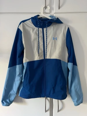 Blå och vit vindjacka från Under Armour storlek s - Snygg vindjacka från Under Armour i blått och vitt med huva och dragkedja. Jackan har meshfoder, justerbara snören vid huvan och logotyp på bröstet. Perfekt för att hålla vinden borta under utomhusaktiviteter. Materialet är lätt och andas.